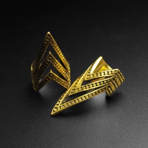May include: Two gold-colored finger rings with a geometric design. Each ring features a series of connected chevrons and small, evenly spaced dots. The rings are displayed against a dark background, highlighting their metallic sheen.