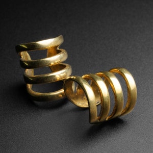 Pode incluir: Duas pulseiras de orelha douradas, cada uma com um design em espiral. Uma pulseira tem quatro faixas e a outra três. As joias são exibidas em uma superfície preta, com a luz refletindo na superfície metálica.