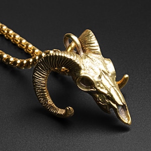 May include: Gold-colored ram skull pendant necklace. The pendant is a detailed sculpture of a ram's skull with curved horns. The chain is a matching gold color and has a rounded, textured design. The necklace is displayed against a dark background.