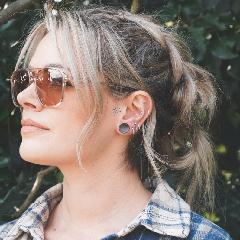 May include: A person wearing clear-framed sunglasses with brown lenses. The person has multiple ear piercings and a dark gray ear plug. A small star tattoo is visible on the face.