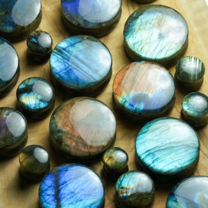 May include: A collection of various sized, round, iridescent gemstone plugs. The plugs are made of labradorite and feature a range of blue, green, and brown hues.
