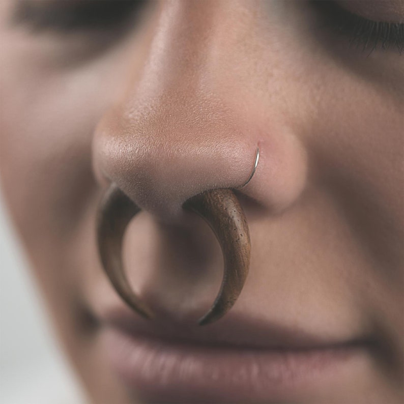 May include: Close-up of a nose with a large, curved wooden nose ring and a small silver nose ring. The wooden ring is a light brown color and has a smooth texture. The silver ring is thin and sits on the nostril.