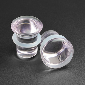 May include: Two clear glass ear gauges with a light purple tint and a white rubber o-ring. The gauges are on a black surface.