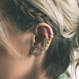 Puede incluir: Primer plano de una oreja adornada con m&uacute;ltiples joyas. Se ve un gran brazalete dorado para la oreja con un dise&ntilde;o en forma de cheur&oacute;n, junto con un peque&ntilde;o pendiente de aro, un pendiente de bot&oacute;n y un tap&oacute;n plateado. Un peque&ntilde;o tatuaje de sol est&aacute; en la mejilla.