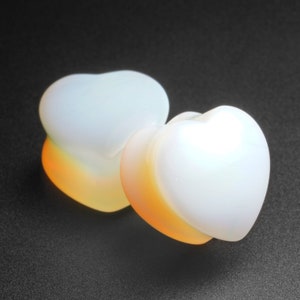 Op de afbeelding: Twee hartvormige plugs, wit aan de bovenkant met een oranje-gele gradiënt aan de onderkant. De plugs zijn glad en ondoorzichtig, tegen een donkere achtergrond. Dit is waarschijnlijk lichaamsjuwelen.