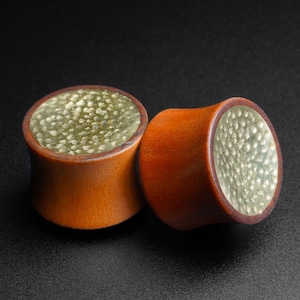 May include: Two wooden ear gauges with a hammered metal inlay. The gauges are brown with a gold-colored metal inlay.