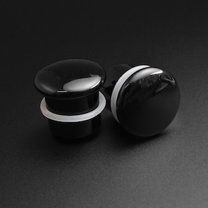May include: Two black ear gauges with a glossy finish and a white silicone ring. The gauges have a rounded, oval shape. The background is a solid black.