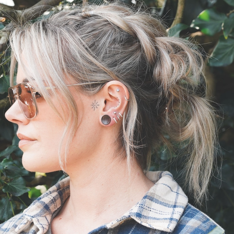 May include: A side profile of a person with blonde hair styled in a loose updo. They are wearing sunglasses and a plaid shirt. Visible ear piercings include multiple rings and a large gauge plug. A small star tattoo is visible.