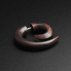 May include: A dark brown, spiral-shaped wooden earring with a silver post. The earring has a smooth, curved design and a natural wood grain appearance. The jewelry is designed to be worn in a stretched earlobe.