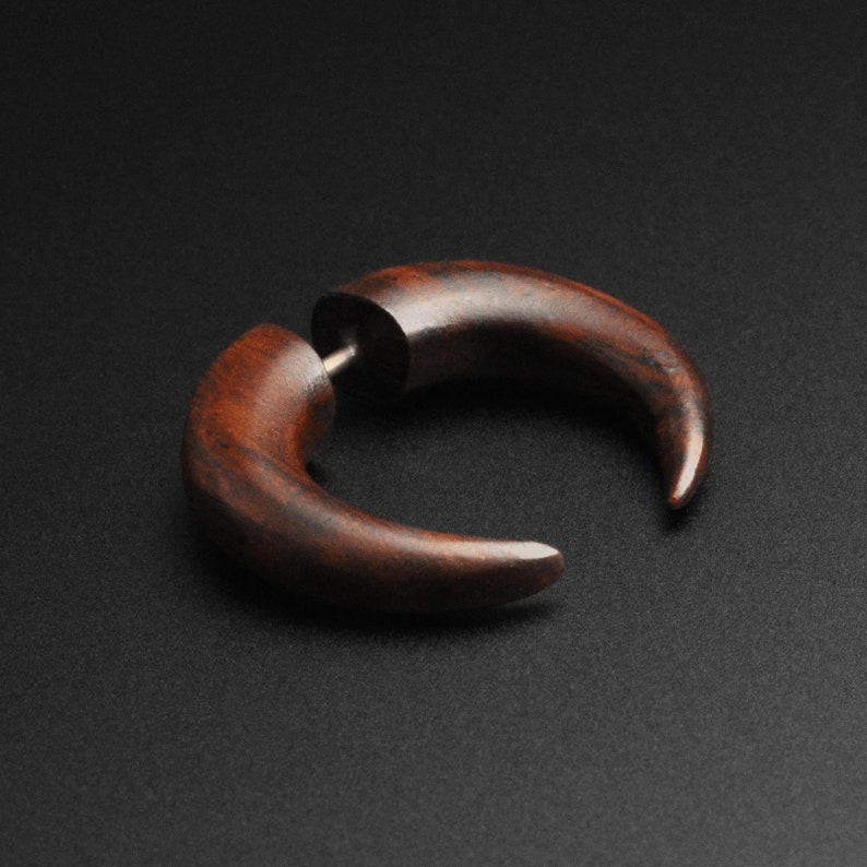 May include: A pair of dark brown wooden earrings in a curved, horn-like shape. The earrings have a smooth, polished finish and a silver post for insertion. The background is a solid black.