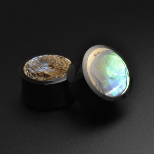 May include: Two black wood ear gauges with abalone shell inlays. The shell inlay on one gauge is a swirling pattern of blue, green, and gold. The other gauge has a more muted brown and gold pattern.