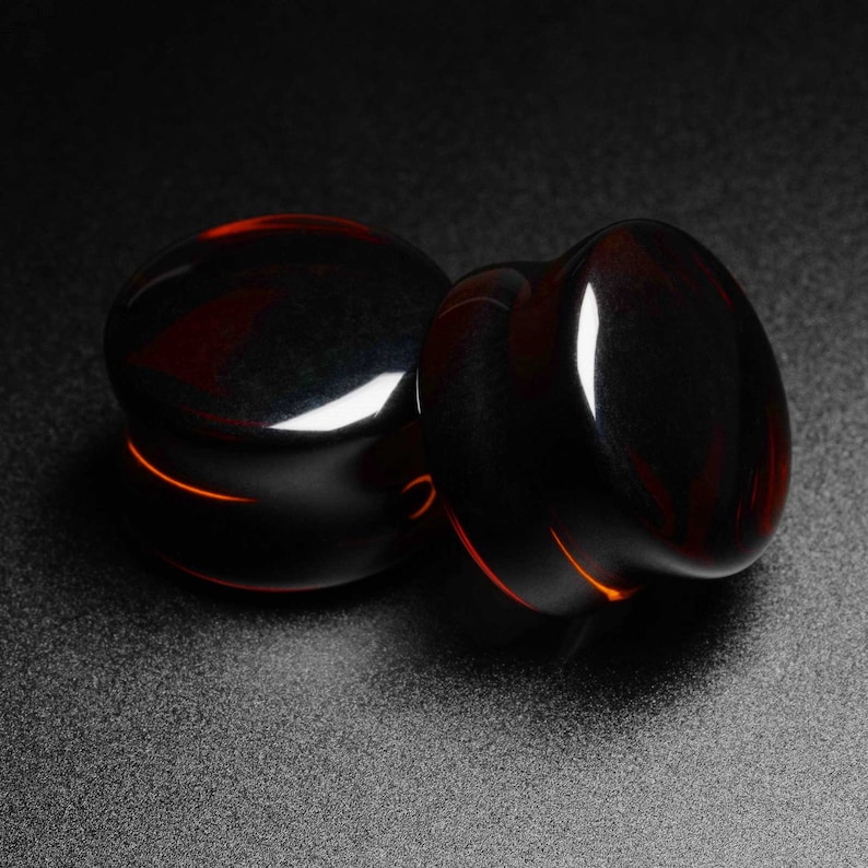 May include: Two black and amber glass ear gauges, one slightly larger than the other, on a black background.