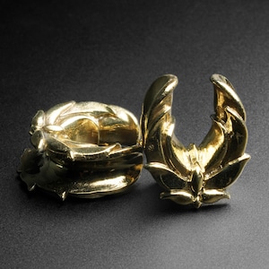 May include: A pair of gold-colored metal ear gauges with intricate floral designs. The gauges are shaped like a ring with a decorative, open-ended design.