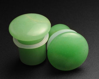 Puede incluir: Dos medidores de oreja de piedra verde, uno con un respaldo de silicona blanco. Los medidores son lisos y tienen un patrón moteado sutil.