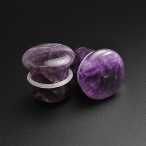 May include: Two amethyst stone ear gauges, one with a white plastic backing. The gauges are a deep purple colour with white streaks throughout.