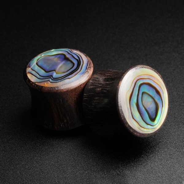 Op de afbeelding: Twee houten oorpluggen met inleg van abalone-schelp. De schelp heeft een draaiende patroon van blauw, groen en wit.