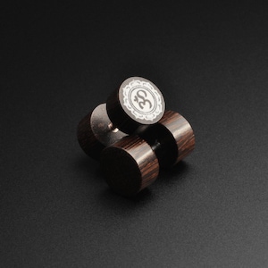 May include: Four dark brown wooden stud earrings. One earring features a white Om symbol within a decorative border. The earrings are cylindrical with a dark wood grain pattern. The background is black.