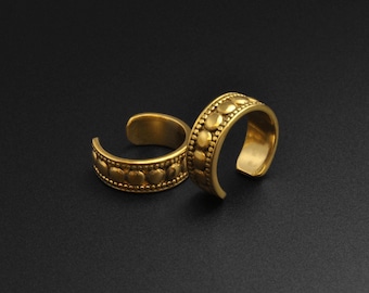 Puede incluir: Dos anillos de dedo del pie dorados con un diseño abierto. Cada anillo presenta una banda adornada con un patrón repetido de pequeños círculos en relieve. Los anillos se muestran sobre un fondo oscuro, resaltando su brillo metálico.