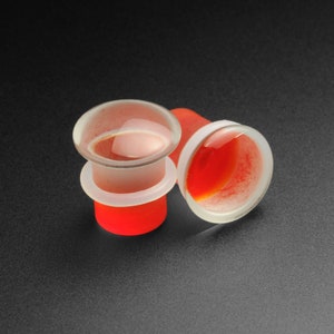 May include: Two clear glass ear gauges with a red interior. The gauges are slightly angled and the one in the foreground is slightly out of focus.