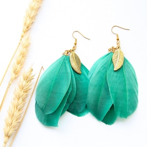 Può includere: Un paio di orecchini di piume color turchese con accenti di foglie dorate. Gli orecchini sono pendenti e hanno uno stile bohémien.