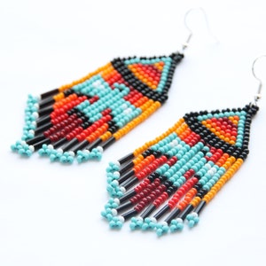 Puede incluir: Un par de pendientes largos de cuentas con un patrón geométrico en turquesa, naranja, rojo, negro y blanco. Los pendientes están hechos con cuentas pequeñas y redondas y tienen ganchos de plata.