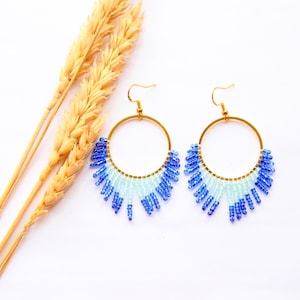 May include: Gold hoop earrings with a blue and white beaded design. The earrings feature a fringe of beads hanging from the bottom. Wheat stalks are in the background. The earrings are a stylish accessory.