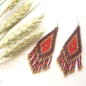 May include: A pair of long, beaded earrings with a geometric design. The earrings are made with red, brown, and gold beads.