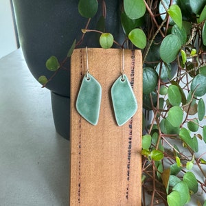 May include: A pair of green ceramic earrings with silver hooks. The earrings are shaped like a teardrop with a pointed bottom and a rounded top. They are displayed on a wooden board with a black and white pattern.