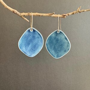 May include: Two blue ceramic earrings with an unusual, irregular form. The earrings are a vibrant blue with a glossy finish and white edges. They hang from delicate gold-coloured ear wires, displayed against a neutral background.