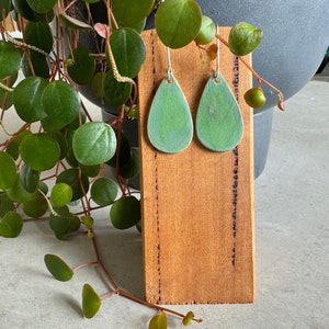 May include: A pair of green ceramic teardrop earrings with silver hooks. The earrings are displayed on a wooden stand.