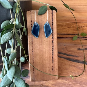 May include: A pair of dark blue, geometric earrings with silver hooks. The earrings are displayed on a wooden block, with green foliage in the background. The earrings have a glossy finish and an unusual, asymmetrical shape.