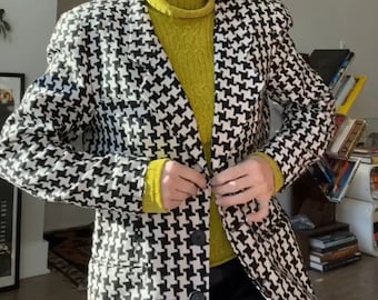 Könnte beinhalten: Eine Person mit grünen Haaren trägt einen schwarz-weißen Hahnentritt-Blazer über einem gelben Rollkragenpullover. Der Blazer hat einen einzigen Knopfverschluss und die Person hält den Knopf mit ihrer rechten Hand.