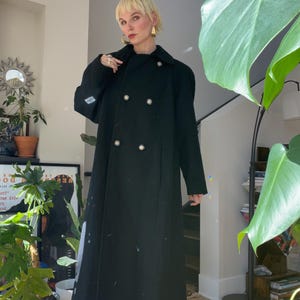 May include: A black double-breasted long coat with pearl buttons. The coat is worn over blue jeans and white platform shoes.