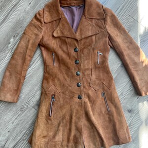 Peut inclure: Un manteau en daim marron avec un col crant&eacute;, une fermeture &agrave; boutons et des poches zipp&eacute;es. Le manteau vintage a une coupe ajust&eacute;e et un aspect doux et textur&eacute;. La doublure est de couleur violet clair.