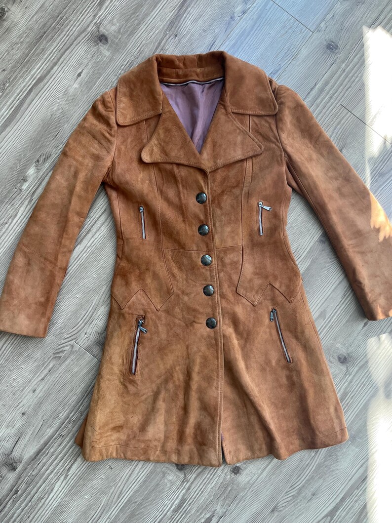 Peut inclure: Un manteau en daim marron avec un col crant&eacute;, une fermeture &agrave; boutons et des poches zipp&eacute;es. Le manteau vintage a une coupe ajust&eacute;e et un aspect doux et textur&eacute;. La doublure est de couleur violet clair.