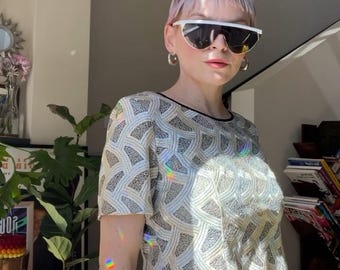 May include: A woman wearing a white, sequined, cropped top with a black trim and a black and white striped skirt. She is wearing white sunglasses and has short, pink hair.