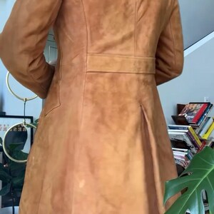 Peut inclure: Un manteau en daim de couleur beige avec un col crant&eacute;, une taille ceintur&eacute;e et une jupe &eacute;vas&eacute;e. Le manteau a un design classique avec une coupe ajust&eacute;e. Le dos pr&eacute;sente un pli pour plus de mouvement. Le manteau est de couleur neutre et chaude.