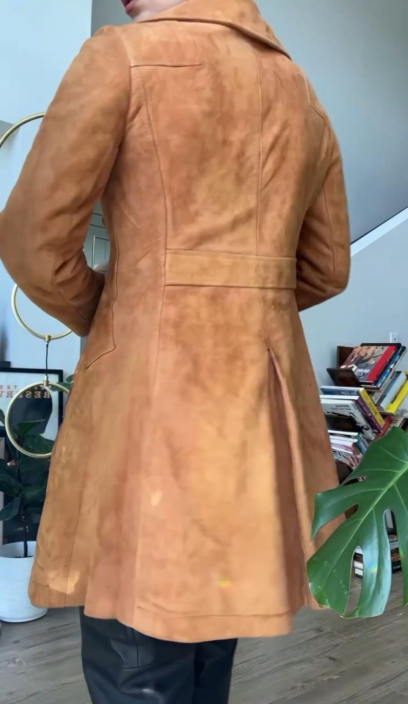 Peut inclure: Un manteau en daim de couleur beige avec un col crant&eacute;, une taille ceintur&eacute;e et une jupe &eacute;vas&eacute;e. Le manteau a un design classique avec une coupe ajust&eacute;e. Le dos pr&eacute;sente un pli pour plus de mouvement. Le manteau est de couleur neutre et chaude.
