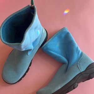 May include: A pair of light blue suede boots with a black sole. The boots have a soft, fleece-like lining and a pull-on design. The boots are displayed on a pink background.