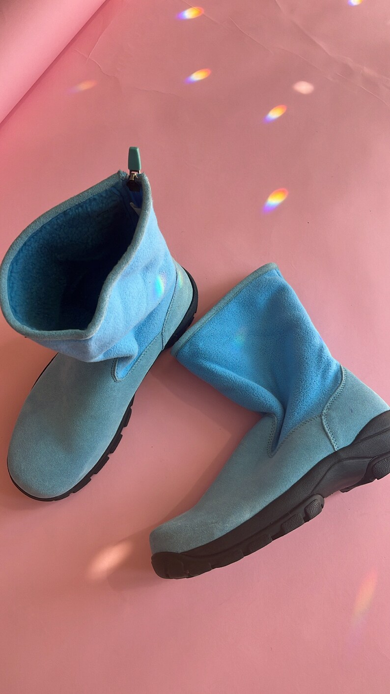 May include: A pair of light blue suede boots with a black sole. The boots have a soft, fleece-like lining and a pull-on design. The boots are displayed on a pink background.