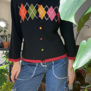 Puede incluir: Un cárdigan negro con ribete rojo y un patrón de diamantes de colores. El cárdigan tiene cierre de botones y está hecho de un tejido suave y de punto.