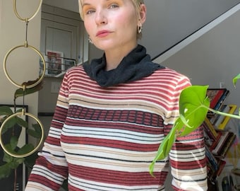 May include: A long-sleeved sweater with horizontal stripes in red, white, and tan. The sweater has a dark gray cowl neck and cuffs. The person is wearing a black skirt. The background includes a window and a decorative gold hanging.