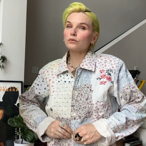 May include: A white patchwork denim jacket with floral and paisley patterns. The jacket has a button-up closure and long sleeves. The person wearing the jacket has bright green hair and is wearing several rings on their fingers.
