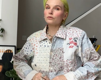 May include: A white patchwork denim jacket with floral and paisley patterns. The jacket has a button-up closure and long sleeves. The person wearing the jacket has bright green hair and is wearing several rings on their fingers.