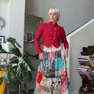 Könnte beinhalten: Eine rote, langärmelige, bauchfreie Jacke mit Stehkragen und goldfarbenen Knöpfen, kombiniert mit einem bodenlangen Rock mit einem mehrfarbigen Patchwork-Design aus verschiedenen Blumen- und geometrischen Mustern sowie einem Kordelzug in der Taille.
