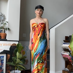 Può includere: Una donna indossa un lungo abito senza spalline, colorato, con una stampa astratta vibrante. L'abito ha un dettaglio annodato sul seno e una silhouette aderente. Indossa un berretto nero e una collana d'argento.