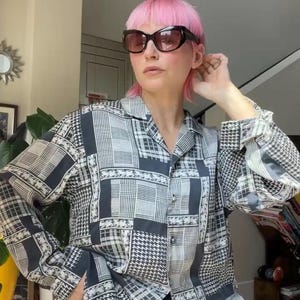 May include: A long-sleeved button-down shirt featuring a black and white patchwork pattern with houndstooth, plaid, and floral motifs. The shirt has a classic collar and is worn with dark-tinted sunglasses with thick black frames.