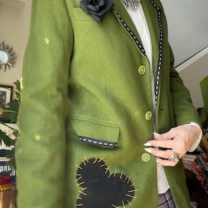 Puede incluir: Una chaqueta verde con ribetes negros y un broche de flor de fieltro negro. La chaqueta tiene un parche de fieltro negro de una carta de picas con costuras amarillas en la parte delantera. La chaqueta se lleva sobre una camisa blanca abotonada y un collar de cadena de plata.