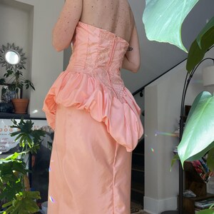 以下が含まれることがあります： ストラップレスのピーチ色のイブニングドレス。ボディスは複雑なビーズ細工とフィットしたシルエットが特徴で、スカートはボリュームのあるフリルペプラムに流れます。ドレスはハイウエストで、光沢のある生地で作られています。