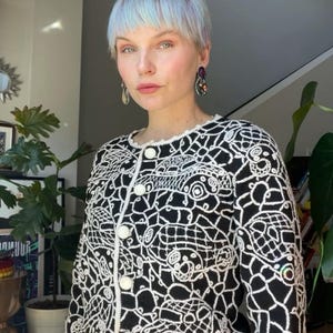 May include: A black and white knit cardigan with a crocheted trim and a unique pattern of white fish and geometric shapes. The cardigan has pearl-like buttons.